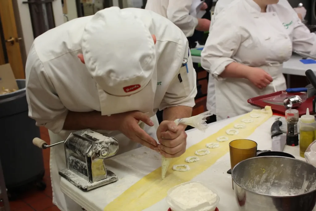 Chef pipes filling onto pasta strips next to a pasta machine in a bustling kitchen.