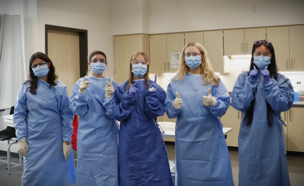 Five people in blue medical gowns, gloves, and masks stand in a clinic, giving thumbs up together.