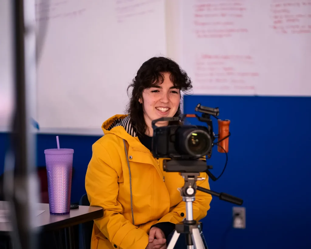 Smiling person in a yellow jacket sits behind a tripod camera in a classroom with notes on the wall.