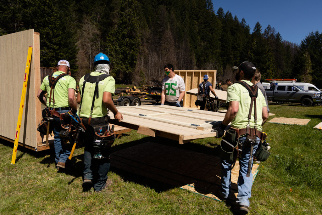 Four workers carry a wall panel while others work nearby on a sunny day with trees and trucks in the background.