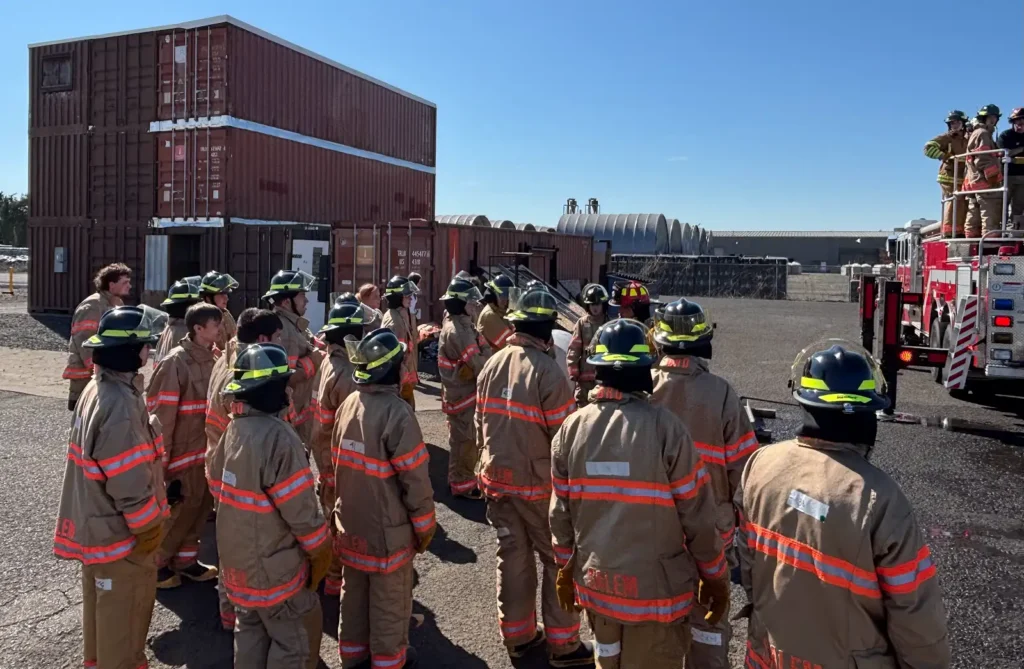 Firefighters in tan gear stand by a fire truck, receiving instructions at an outdoor training area.