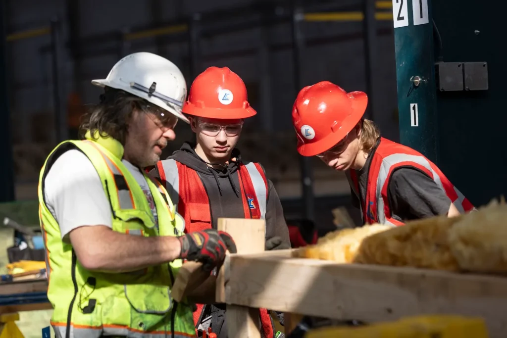 Three construction workers in safety gear inspect a wooden beam together at a sunlit construction site.