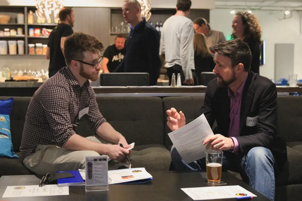 Two men with papers talk on a couch amid a busy networking event in a modern lounge.