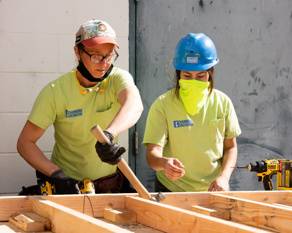 Construction workers in safety gear assemble a wooden frame outdoors under bright sunlight.