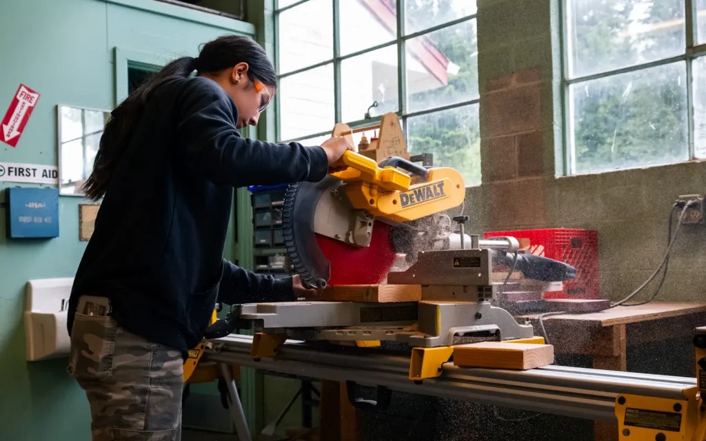Person cuts wood with a DeWalt miter saw in a bright workshop featuring large windows and a first aid kit.