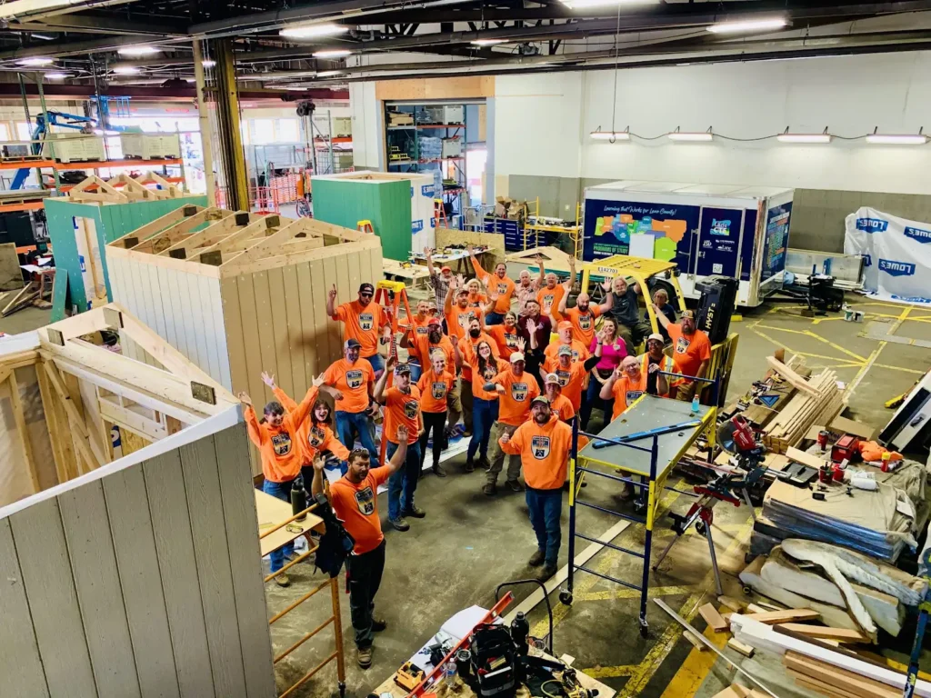 People in orange shirts wave together inside a bustling workshop filled with wooden structures and tools.