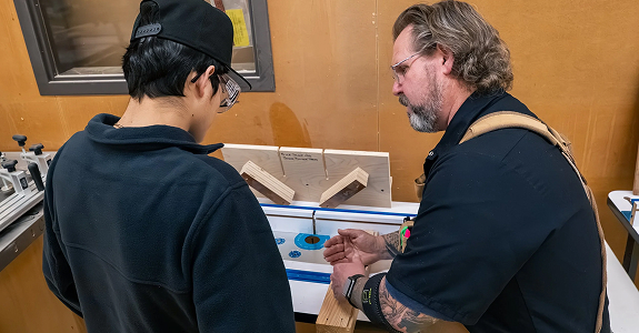 Instructor guides student using woodworking tools at a table as both wear safety glasses.