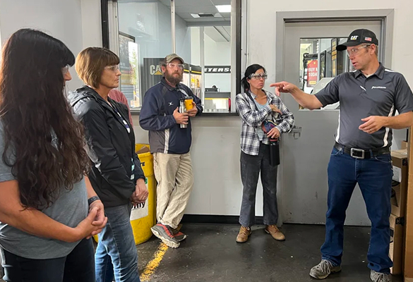 Five people stand indoors while a man in a gray shirt speaks and gestures with his hand.