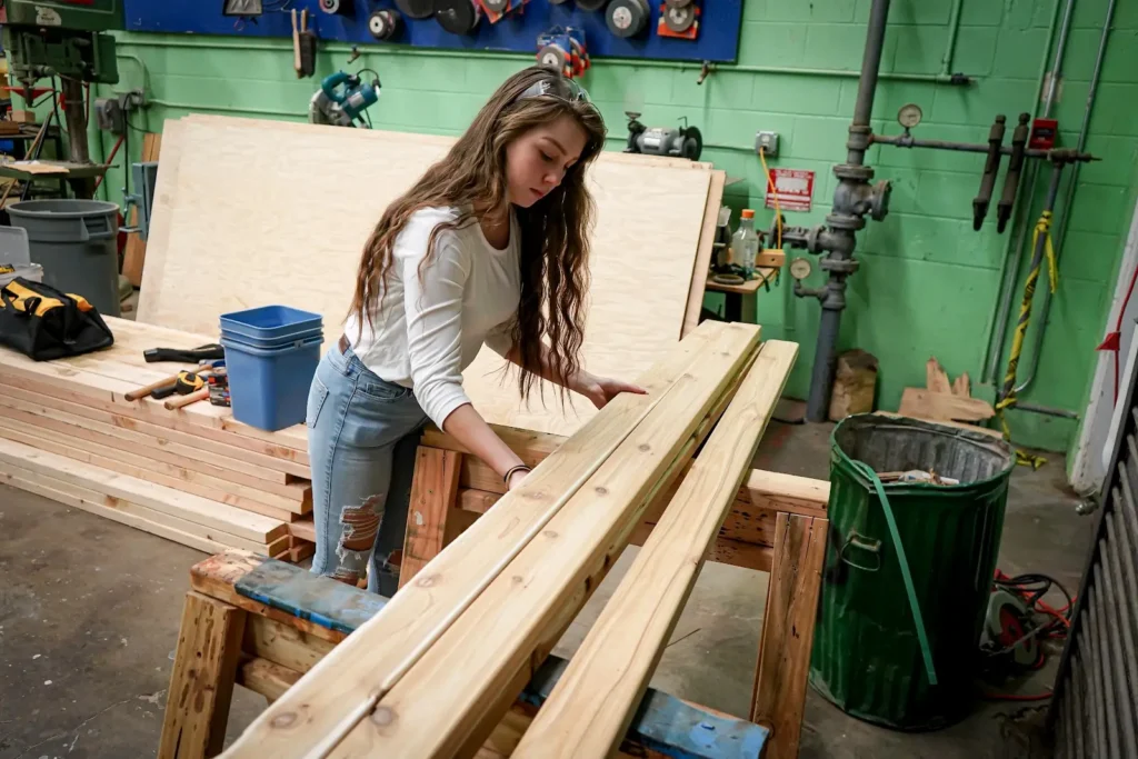 A woman examines wooden planks in a workshop filled with tools and various materials.