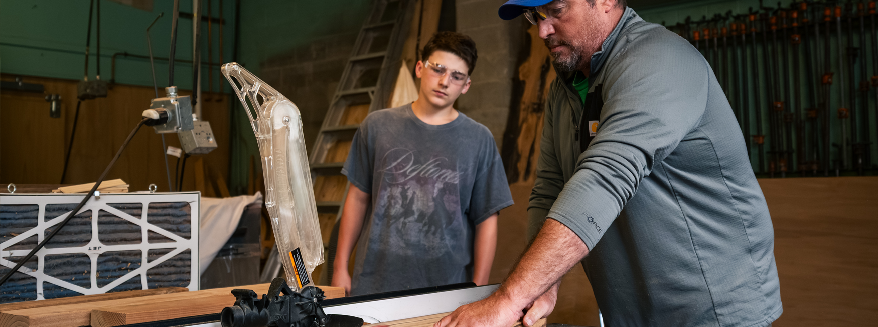 A man operates a table saw on wood as a boy watches closely in a busy workshop.
