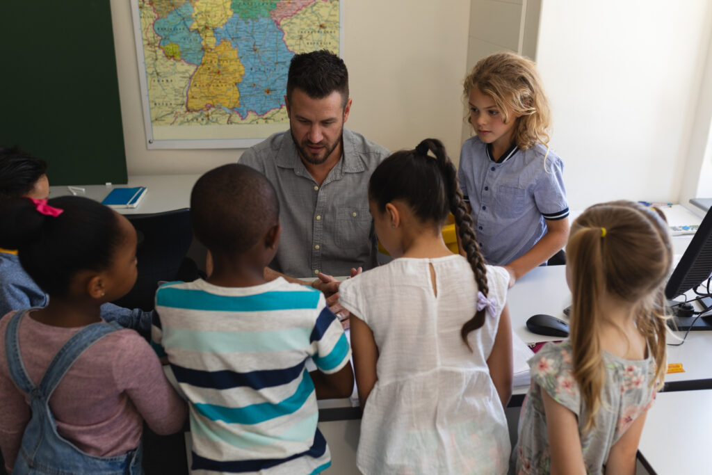 A teacher at a desk speaks as several young students gather around, listening with focused attention.